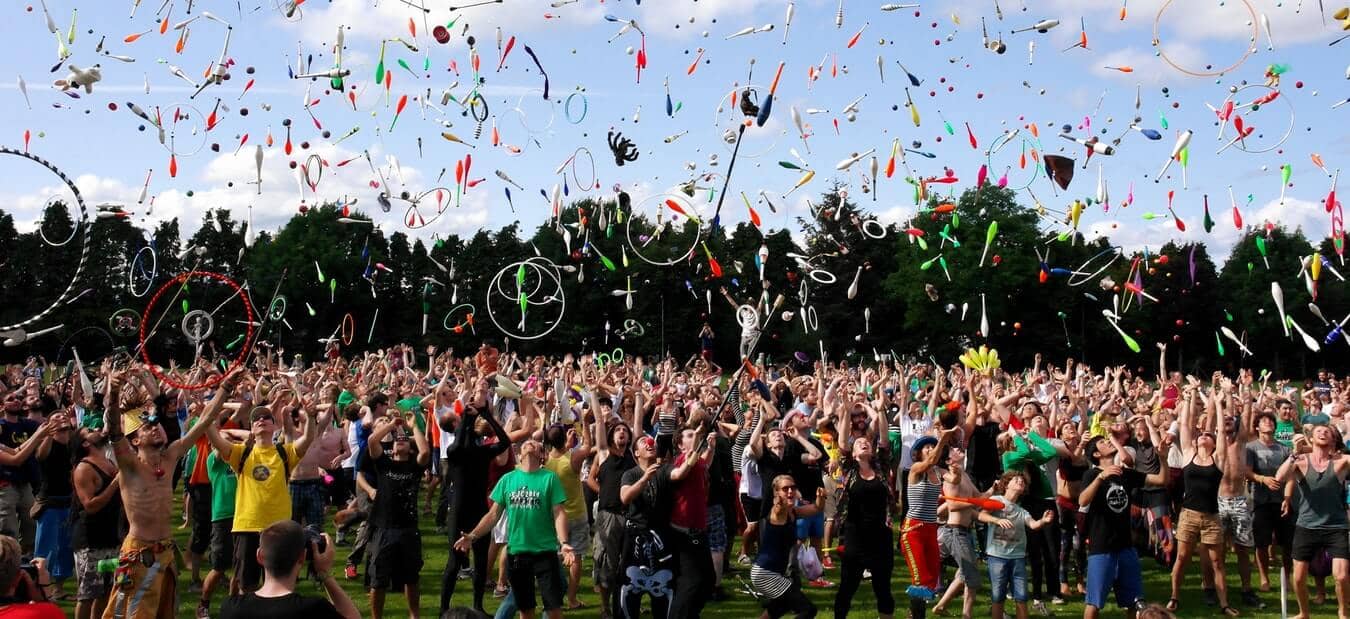 A large crowd outdoors throwing juggling clubs and rings into the air against a blue sky and tree backdrop.
