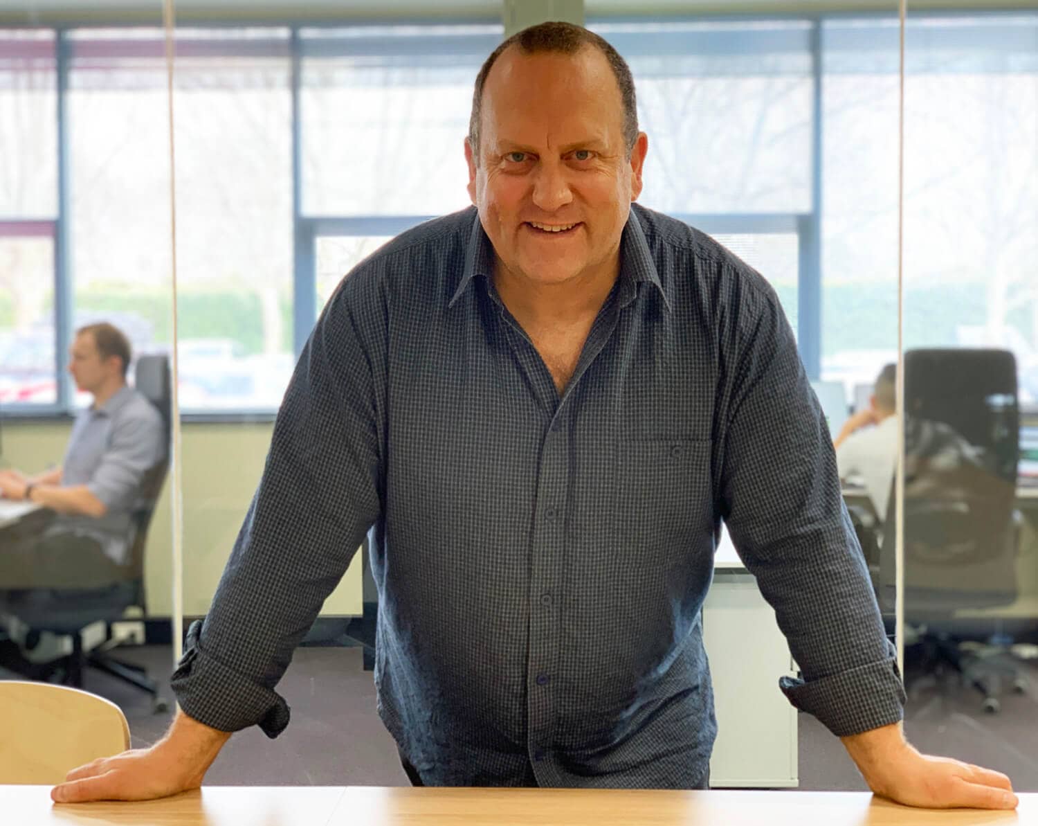A middle-aged man in a blue checkered shirt leaning on a desk, smiling, with two people working in an office behind him.