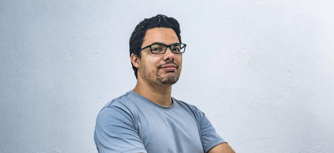 Man with glasses and short curly black hair, wearing a blue t-shirt, standing confidently against a plain white wall.