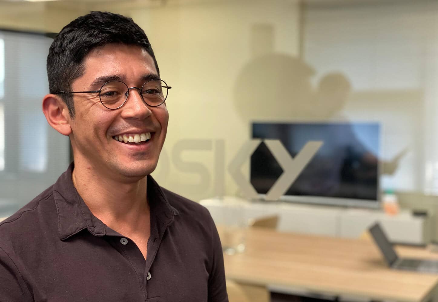 Smiling man wearing glasses and a dark shirt in a modern office with a blurred screen and logo in the background.