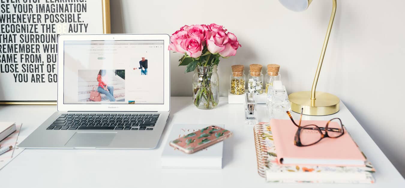 White workspace with a laptop, pink roses in a jar, stationery, a gold lamp, and glasses resting on notebooks.