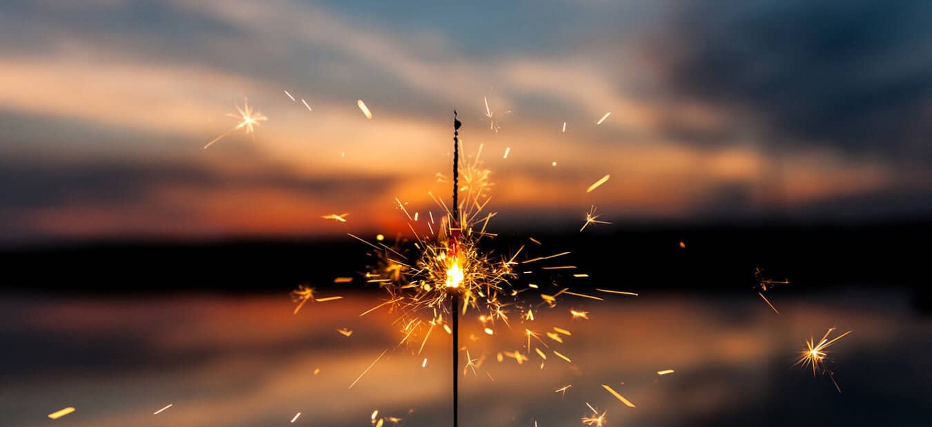 A lit sparkler glowing with bright sparks against a blurred sunset sky and calm water background.