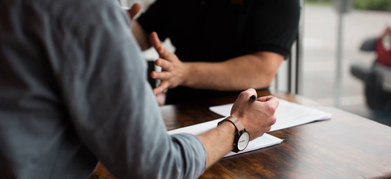 Two people at a wooden table discussing documents, one holding a pen and gesturing with hands.
