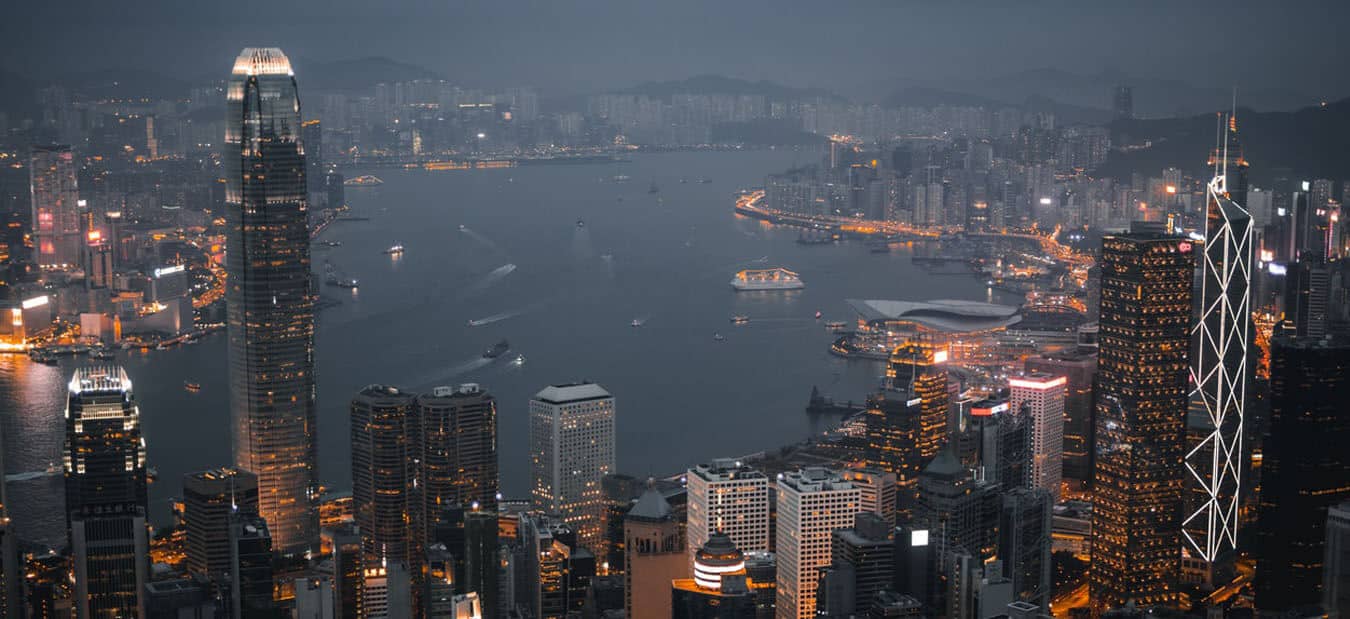 Nighttime aerial view of a vibrant city skyline with illuminated skyscrapers and a busy harbor with boats.