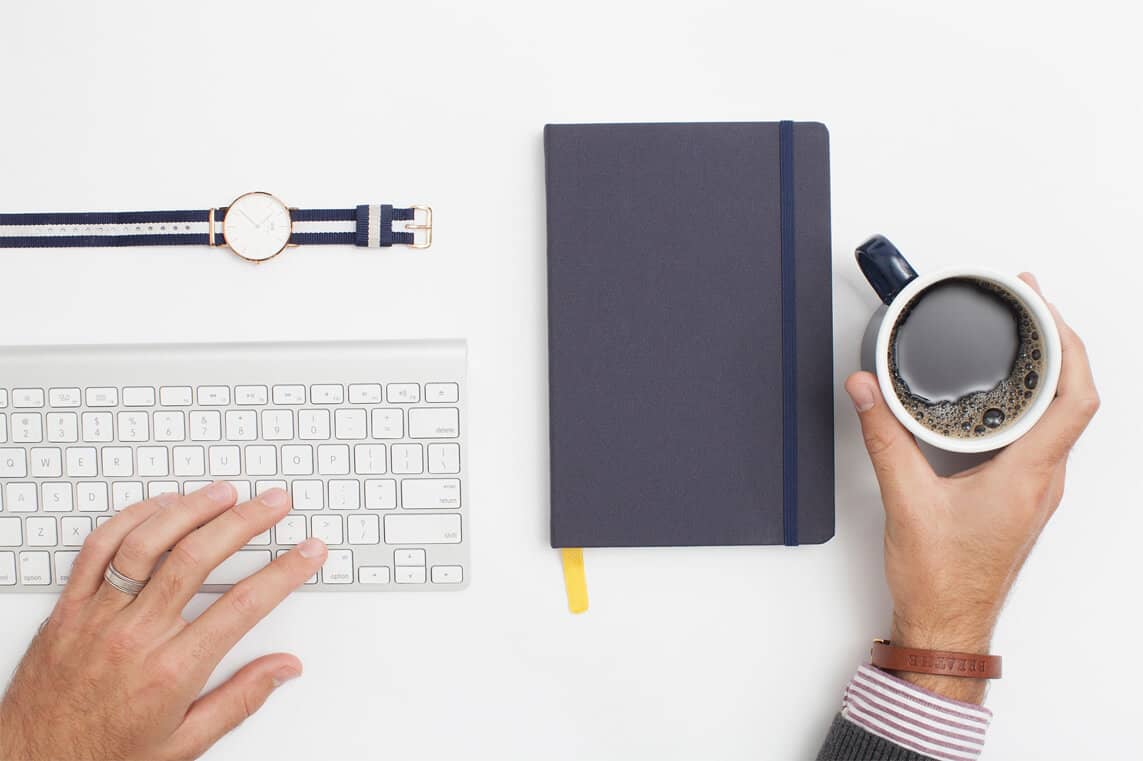 Hands typing on a white keyboard and holding a coffee cup next to a closed navy notebook and striped watch on a white desk.
