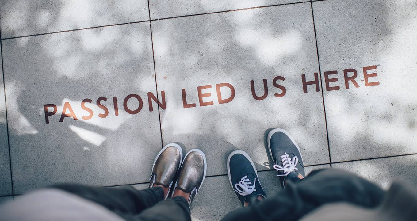 Two pairs of feet standing near sidewalk text that reads "PASSION LED US HERE" with dappled sunlight.
