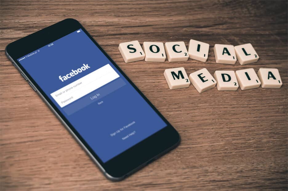 Smartphone displaying Facebook login screen beside Scrabble tiles spelling "SOCIAL MEDIA" on wooden surface.