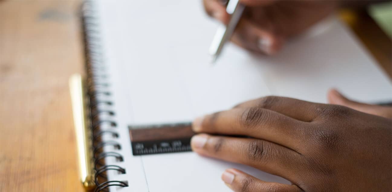 Close-up of a person using a small ruler and pen to draw precise lines on a spiral notebook with a wooden table in the background.