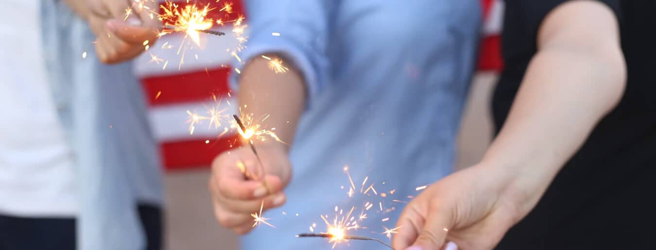 People holding sparklers with sparks glowing, with a blurred American flag in the background.