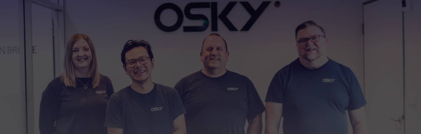 Four smiling coworkers wearing matching black OSKY t-shirts standing side by side against a white office wall with the OSKY logo.