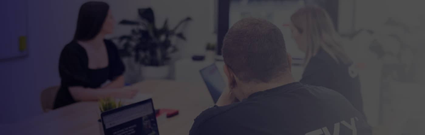 Three people in a meeting room working on laptops, with a plant and whiteboard in the background.