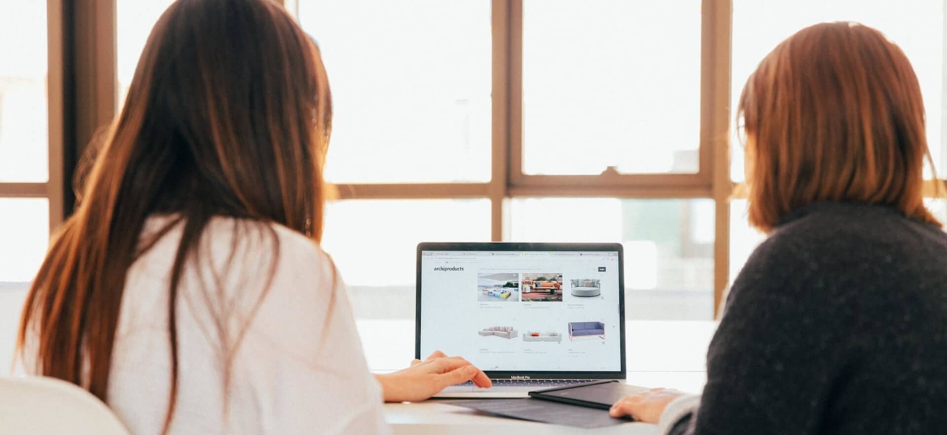 Two women discussing furniture options on a laptop during requirement gathering and discovery phase in an agile setting.