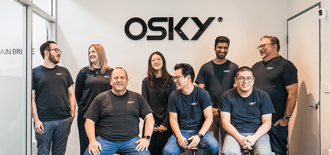 Group of eight people in matching black Osky shirts smiling and posing in front of a wall logo.