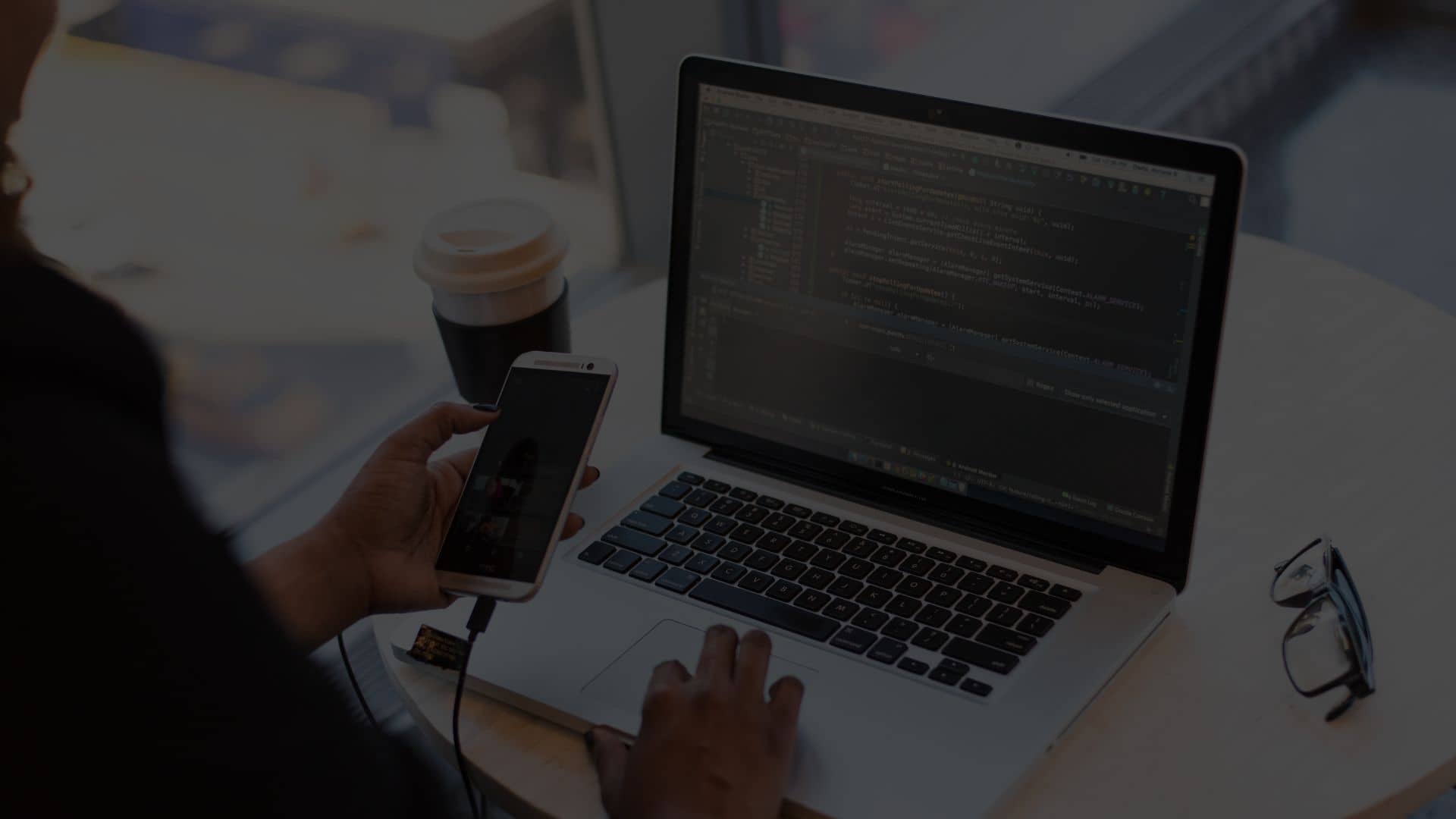 Person conducting web testing on laptop and smartphone at round table with code on screen, coffee cup, and glasses nearby.