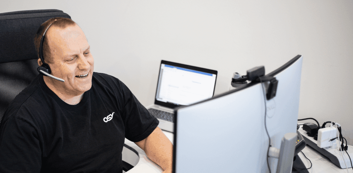 Man wearing headset, smiling while working at a desk with dual monitors and a laptop.