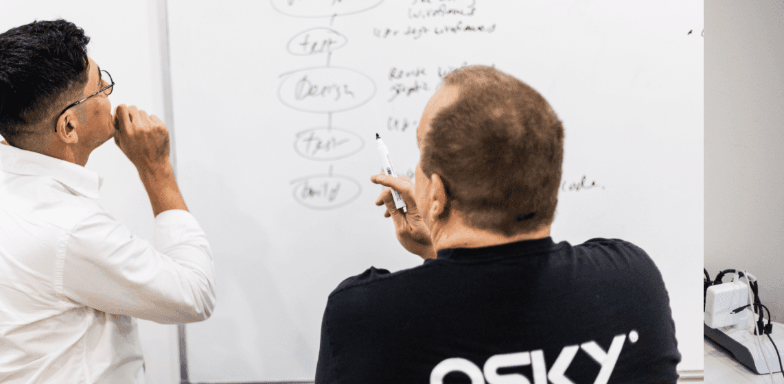 Two men discuss and write on a whiteboard with flowcharts and notes during a meeting.