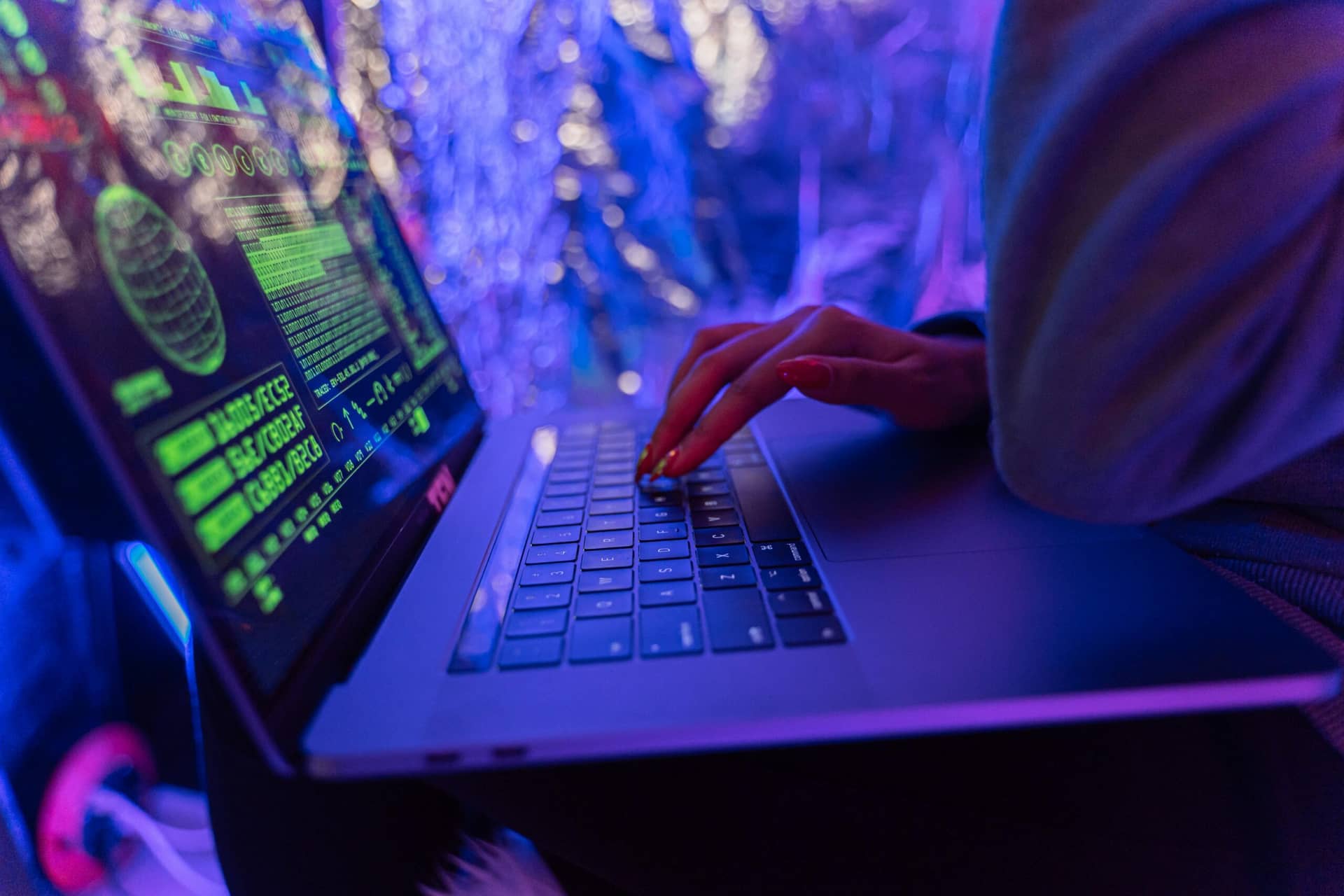 Person typing on laptop with green code on screen in dim purple-lit room, showcasing cybersecurity essentials for nonprofi...