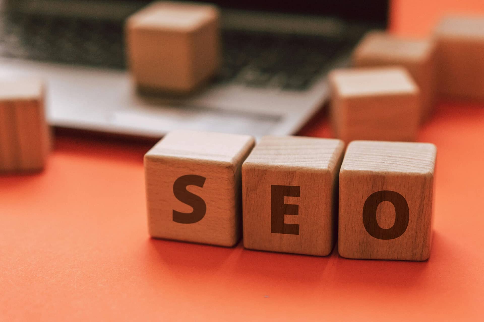 Wooden blocks spell "SEO" on orange surface, blurred laptop keyboard in background - SEO website audit report concept.