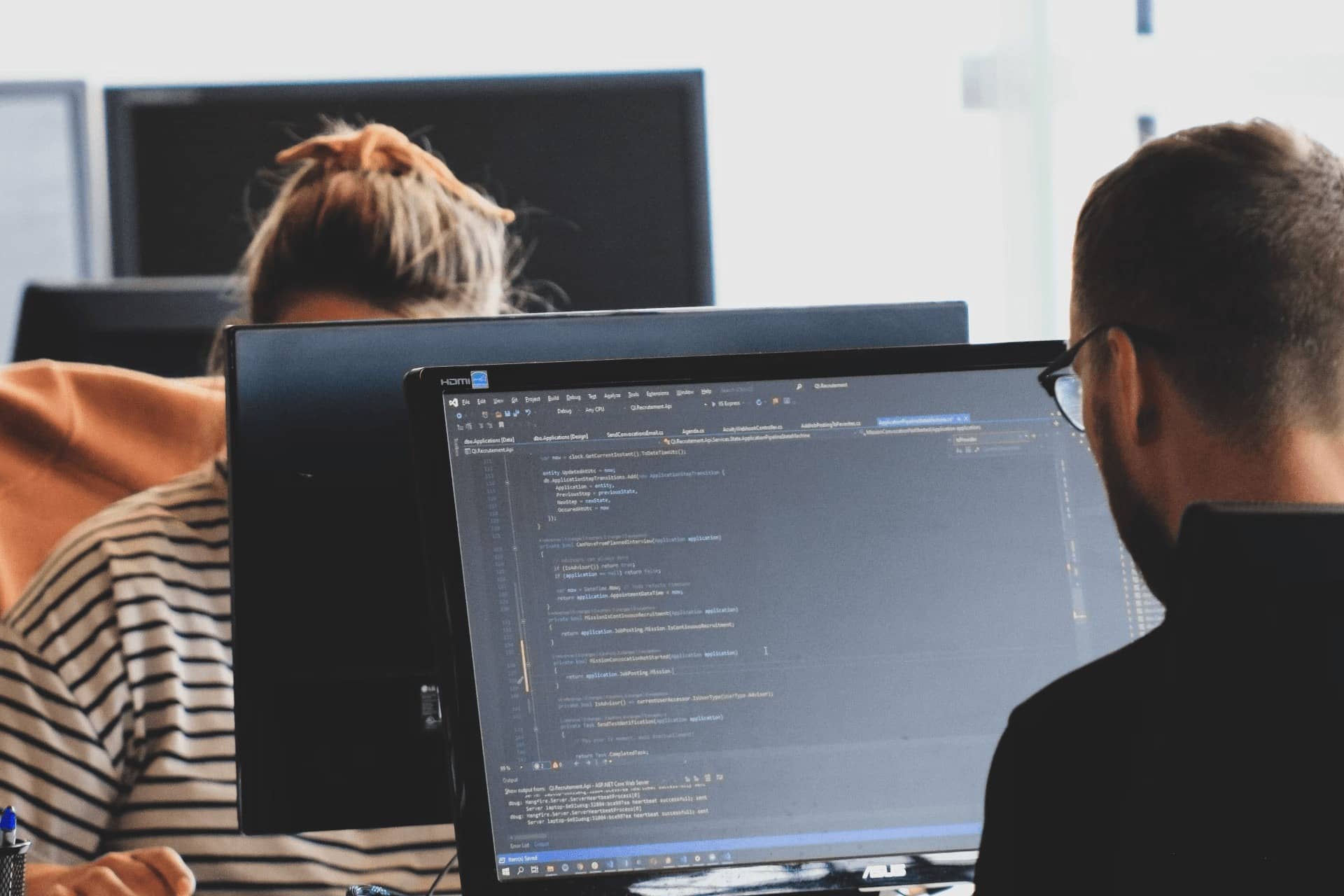 Two people working at computers, one screen displaying programming code in a bright office environment.