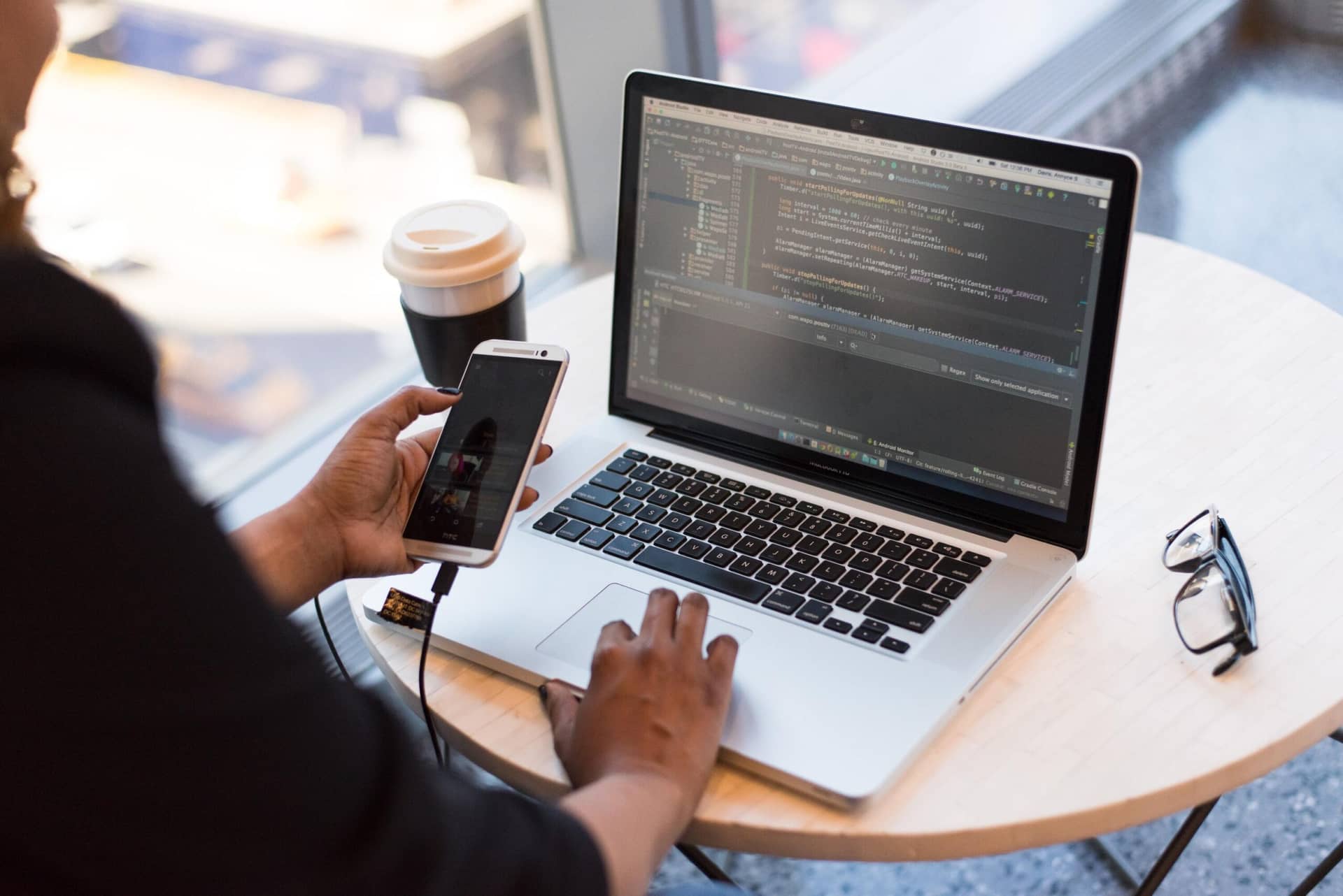 Person working on laptop with coding software, holding connected smartphone, with coffee cup and glasses on round table.
