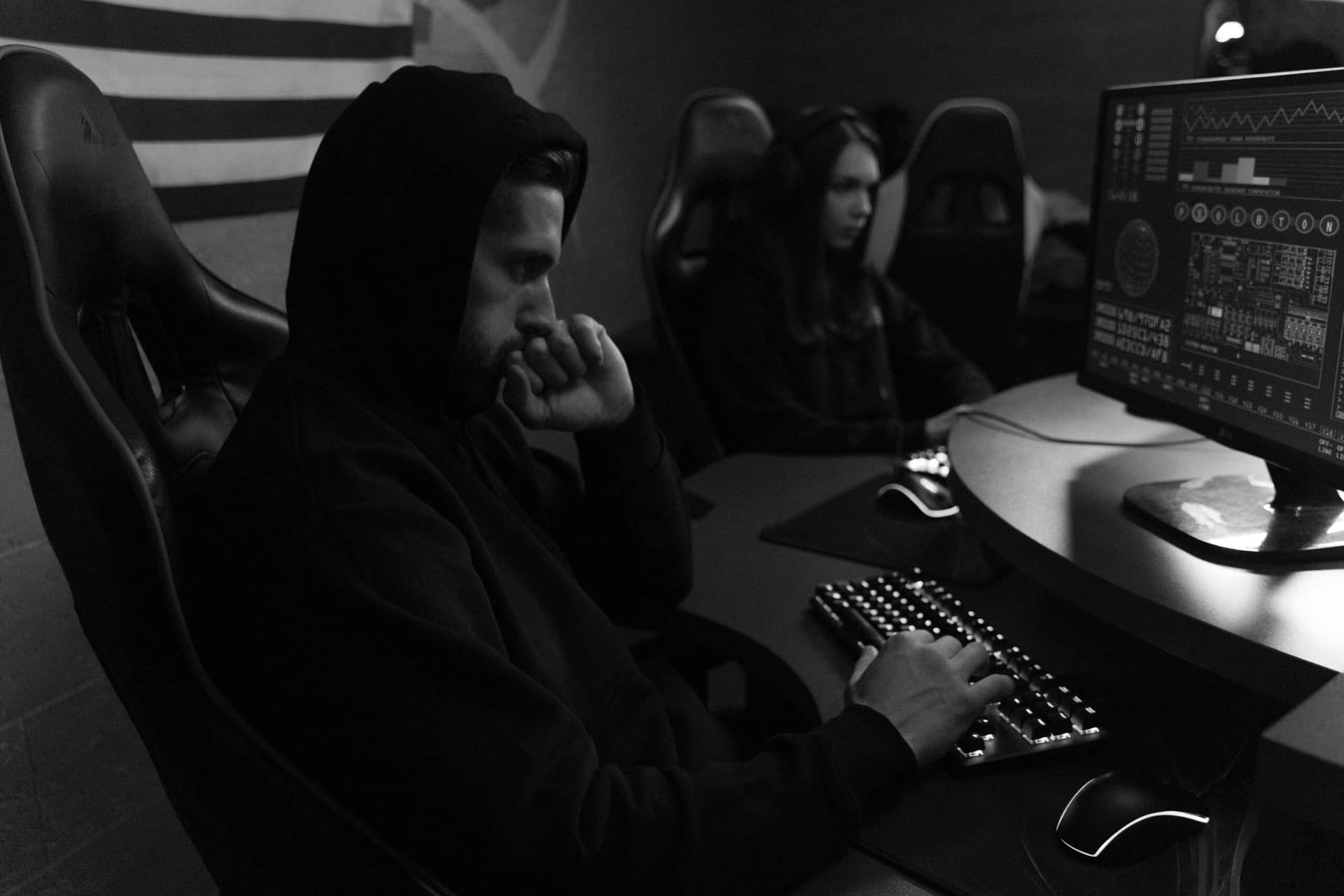 Two cybersecurity experts in hoodies working on computers with web application firewall code in dimly lit room.